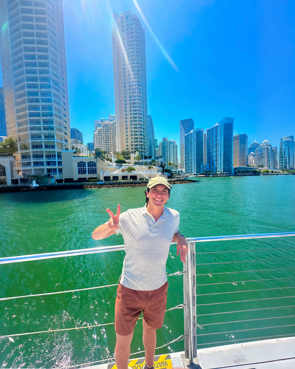 Matthew Vargas smiling on a boat, flashing a peace sign with turquoise water and a city skyline behind him
